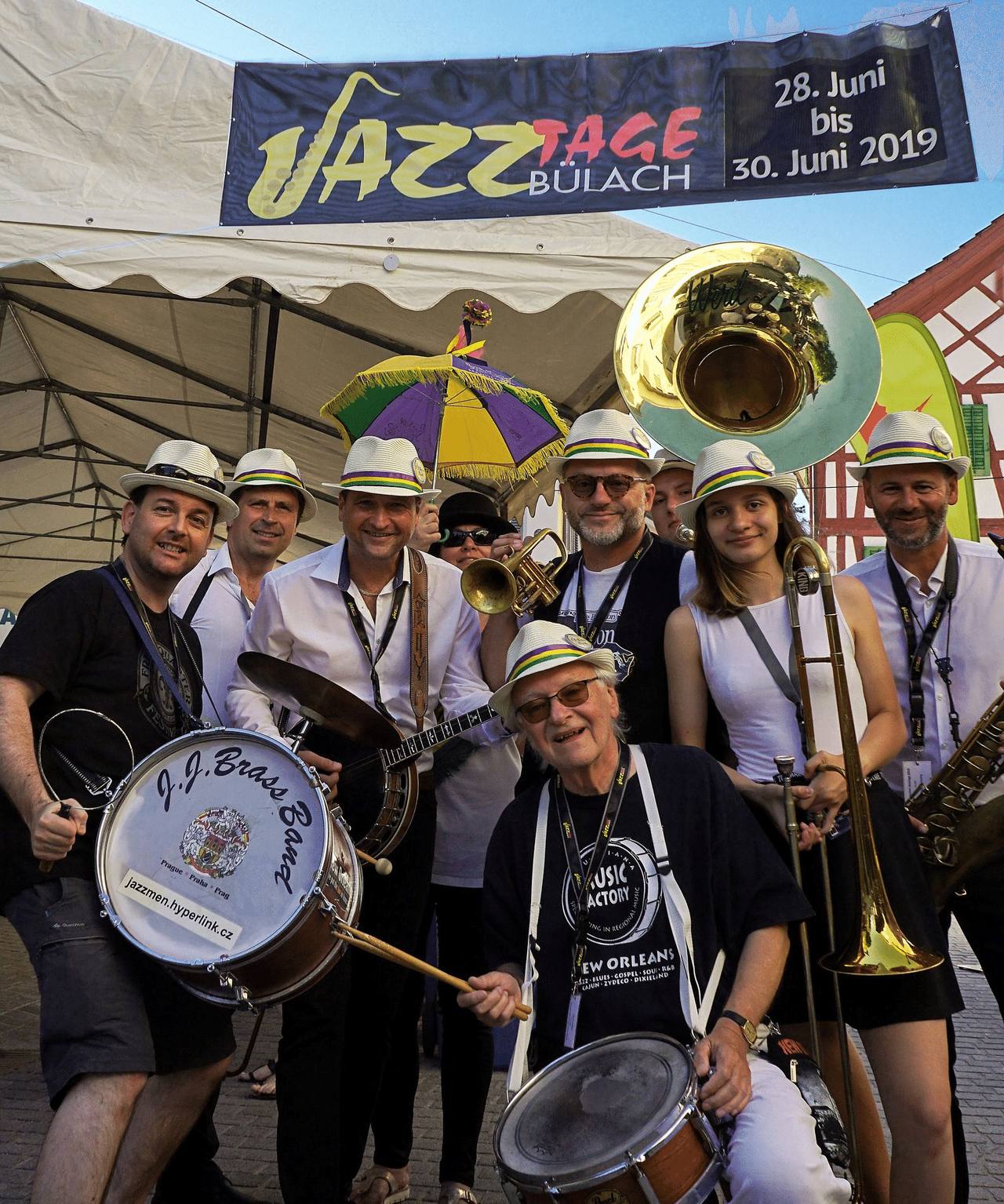 Členové J.J. Brass Band v bílých kloboucích s tubou a bubnem, jeden z nich s pozounem.