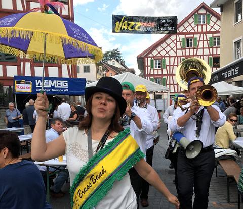 Zpěvačka J.J. Brass Band v cylindru a s šerpou vede průvod hudebníků ulicí městečka.