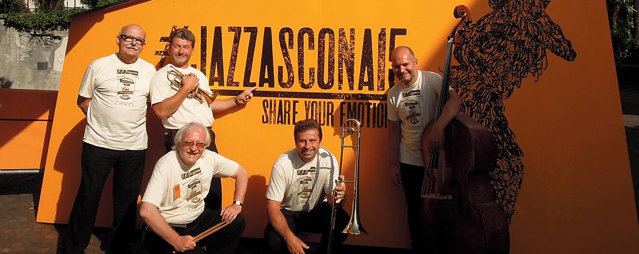 Šest hudebníků v tričkách kapely pózuje před oranžovým nápisem festivalu Jazz Ascona.