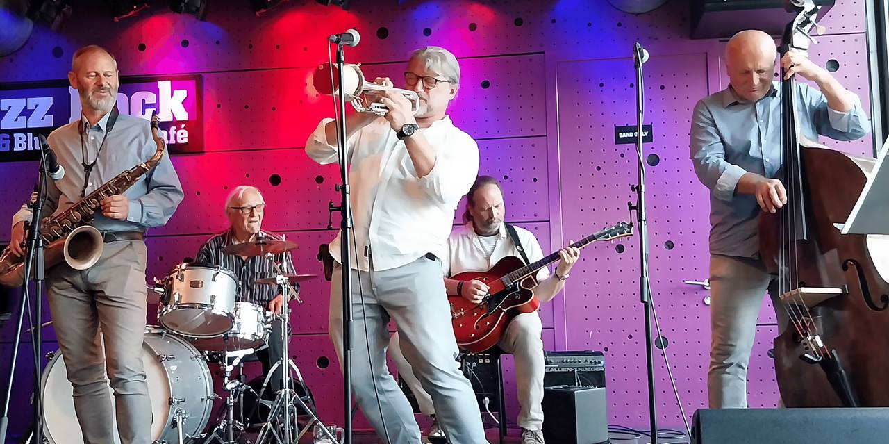 Koncert J.J. Jazzmen v klubu Jazz Dock: trumpetista, saxofonista, bubeník, kytarista a kontrabasista na barevně nasvíceném pódiu.