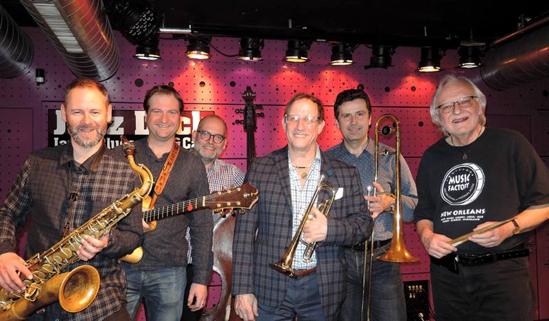 Členové J.J. Jazzmen s hostem, držitelem ceny Grammy Kevinem Rayem Clarkem, na pódiu se saxofonem, kytarou a pozounem.